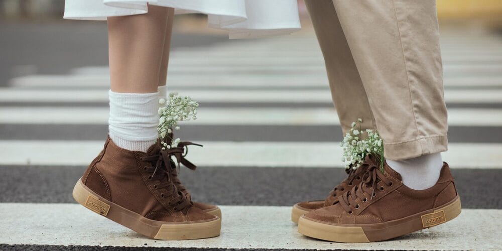 Two people standing face to face on a crosswalk, wearing flexible brown canvas sneakers with flowers tucked inside their socks.