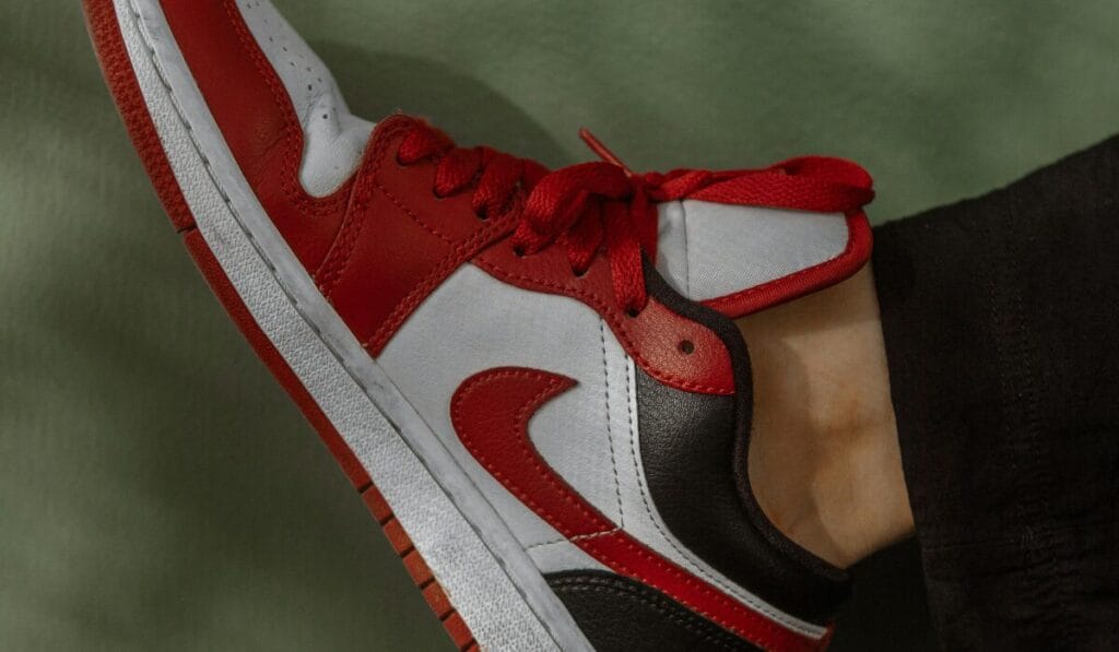 Red, white, and black athletic sneaker with red laces, worn by a person dressed in black pants, lifted slightly against a green background.