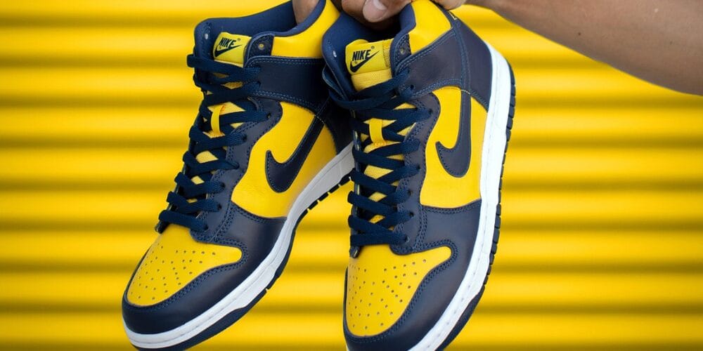 Yellow and navy blue Nike sneakers with white soles held by hand against a yellow background.