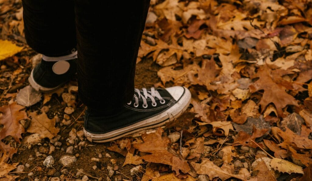 Person wearing black canvas walking shoes on a dry, leaf-covered trail in autumn.