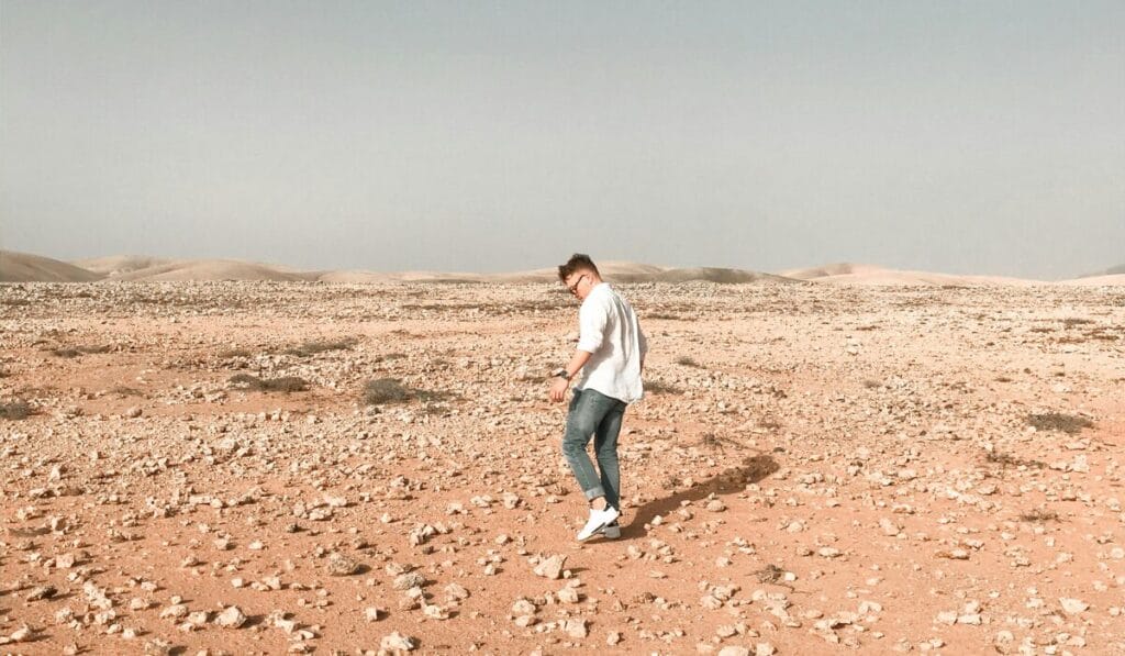 A man walking on a dry, rocky desert terrain wearing casual shoes and jeans.