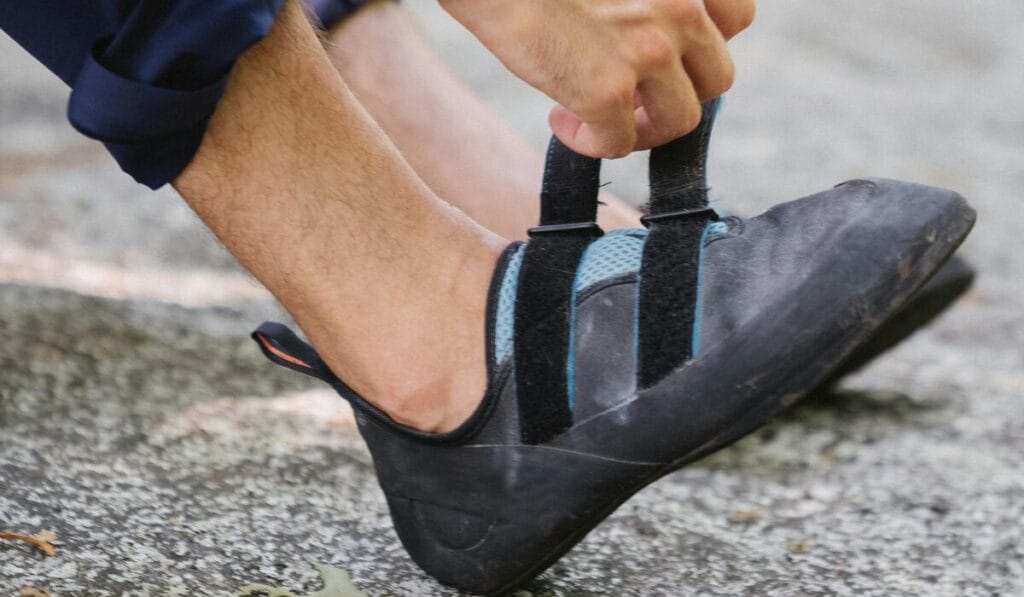 Close-up of a person wearing black walking shoes with Velcro straps, tightening the straps while barefoot on an outdoor surface.
