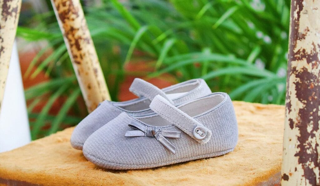 Pair of light gray baby shoes with straps and decorative bows placed on a rustic surface.