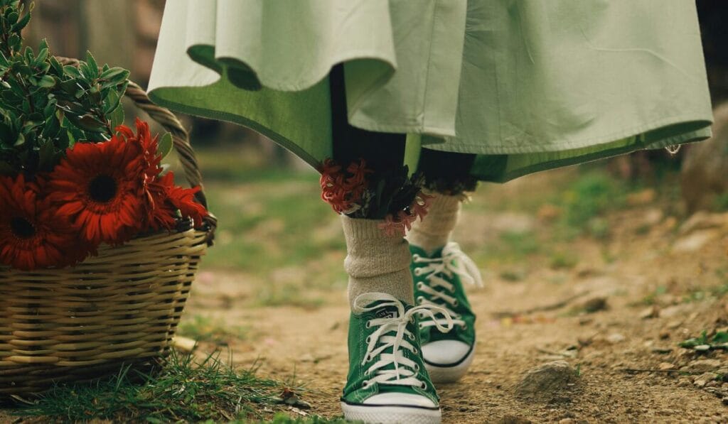 Green canvas walking shoes styled with a light green dress, worn outdoors on a dirt path with a basket of red flowers.