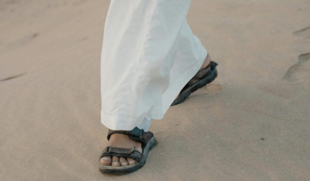 Black walking sandals worn with a white traditional dress, walking on sandy ground.