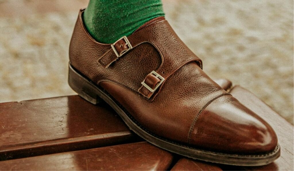 Close-up of a brown leather double monk strap shoe with green socks, resting on a wooden bench.