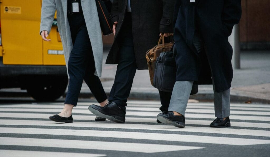 A group of people in dark coats wearing comfortable walking shoes crossing a city street on a crosswalk.