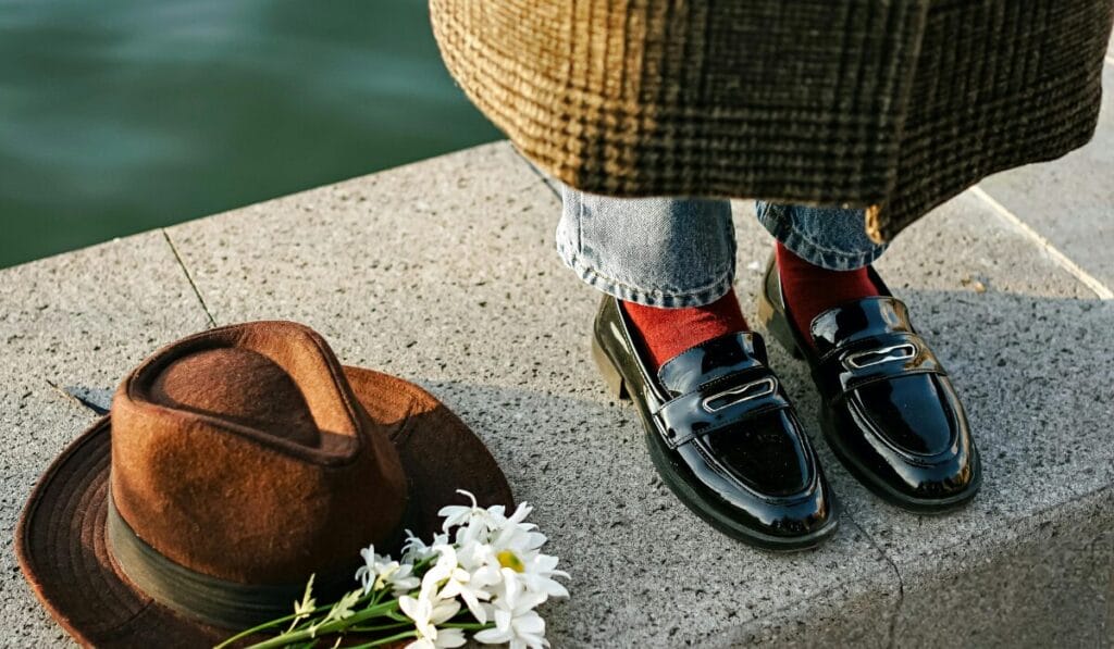 Person wearing black leather loafers, red socks, and blue jeans standing near a brown hat and white flowers on a concrete surface.