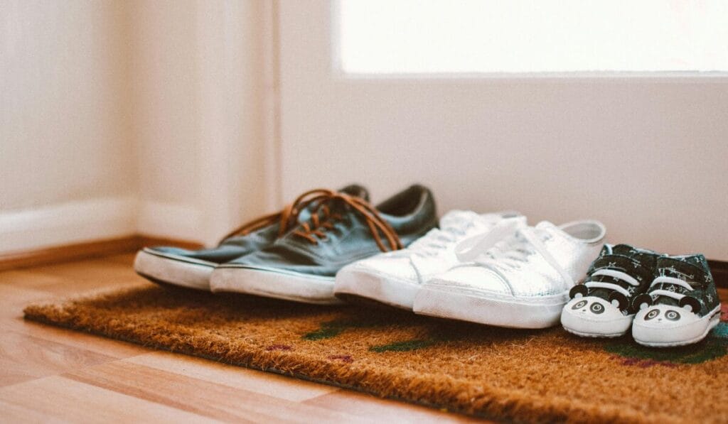 Three pairs of shoes placed on a doormat — one black with brown laces, one plain white, and one tiny black-and-white panda design shoes for a baby.