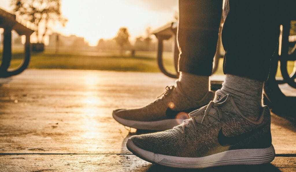 Close-up of a person wearing running shoes and socks, standing outdoors on a wooden floor during sunset.
