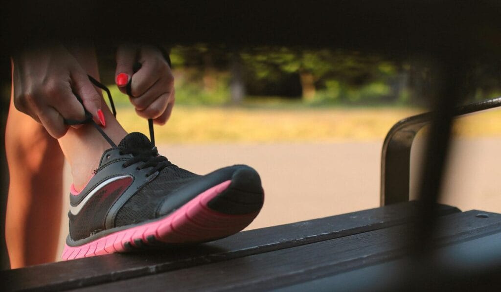 Close-up of a person tying the laces of black and pink running shoes while sitting on a bench outdoors.
