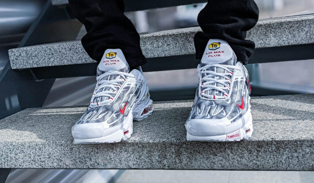 Person wearing Nike Air Max Plus Tuned Air sneakers with red swoosh design, sitting on concrete stairs.