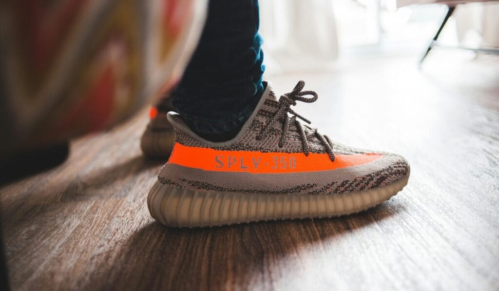 Close-up of a person wearing Adidas Yeezy Boost 350 sneakers with orange stripe and 'SPLY-350' text, standing on wooden floor.