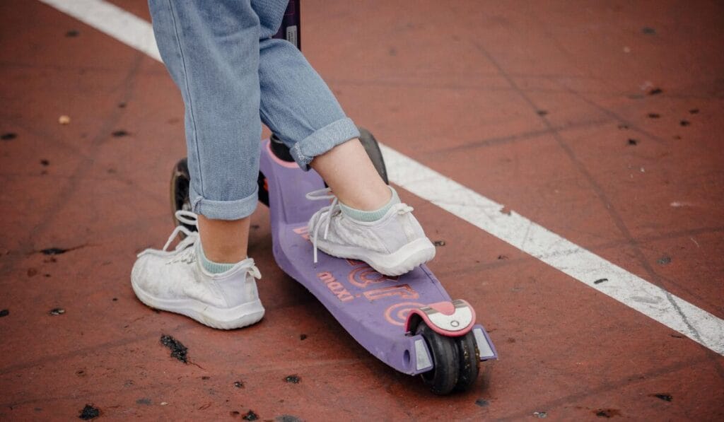 Child wearing white sneakers riding a purple kick scooter on a red sports court.