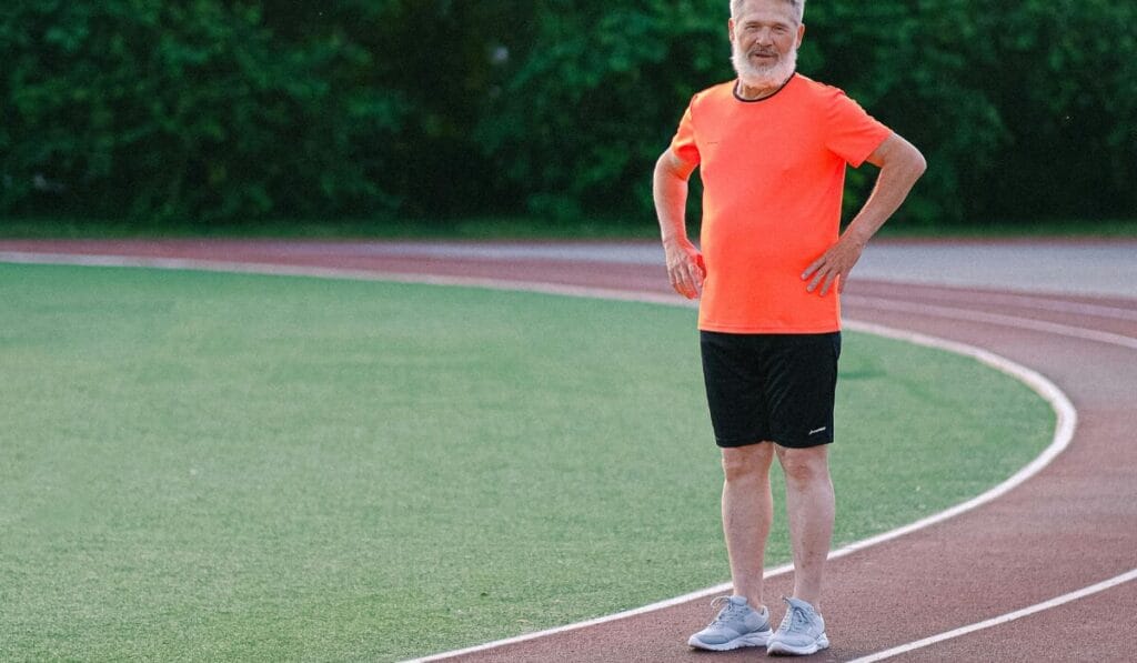 Older man in bright orange sports shirt and black shorts standing on a running track wearing gray walking shoes.