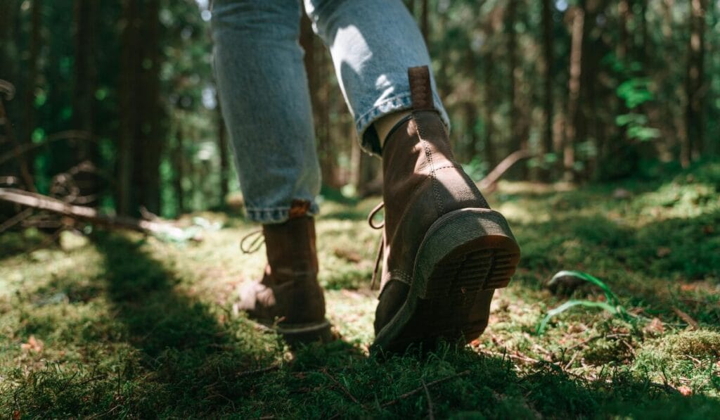 Close-up of a person wearing brown hiking boots walking through a forest trail with moss-covered ground.