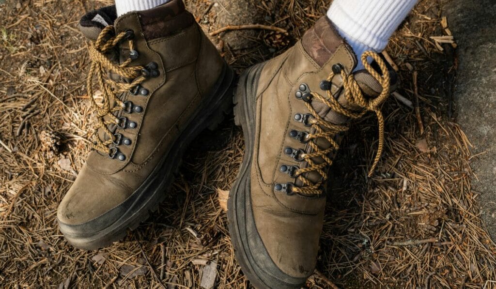 Pair of rugged brown hiking boots with yellow laces worn with white socks, standing on a forest floor covered in pine needles.