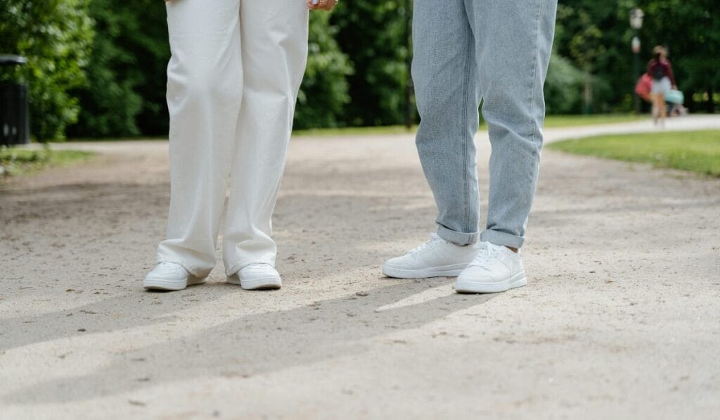 Two people standing on a park pathway wearing white sneakers, one dressed in white pants and the other in light blue jeans.