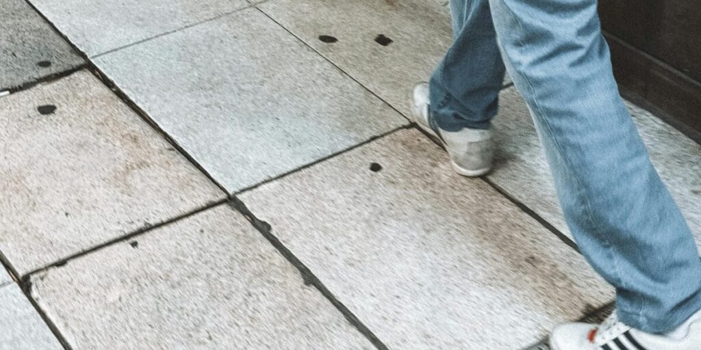 A person wearing blue jeans and white sneakers walking on a tiled sidewalk.